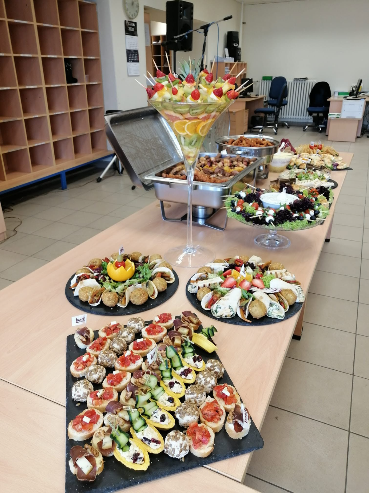 Dugi kancelarijski stol postavljen za koktel — voće u velikoj čaši, topla jela u posudama, platoi kanapea i rolnica — Lunch Paket Catering