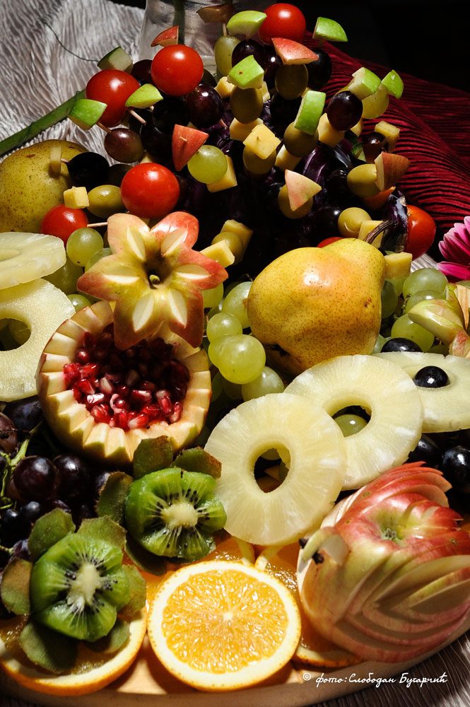 Voćna postavka — kolutovi ananasa, kivi, grožđe, kruške, nar u izrezanoj dinji i ražnjići od voća
